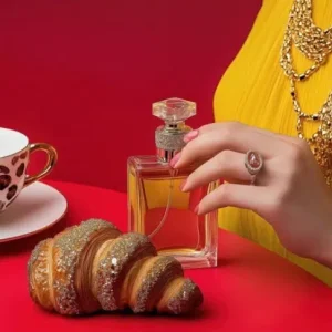 Une main tenant un parfum près d'un croissant et d'une tasse de café.